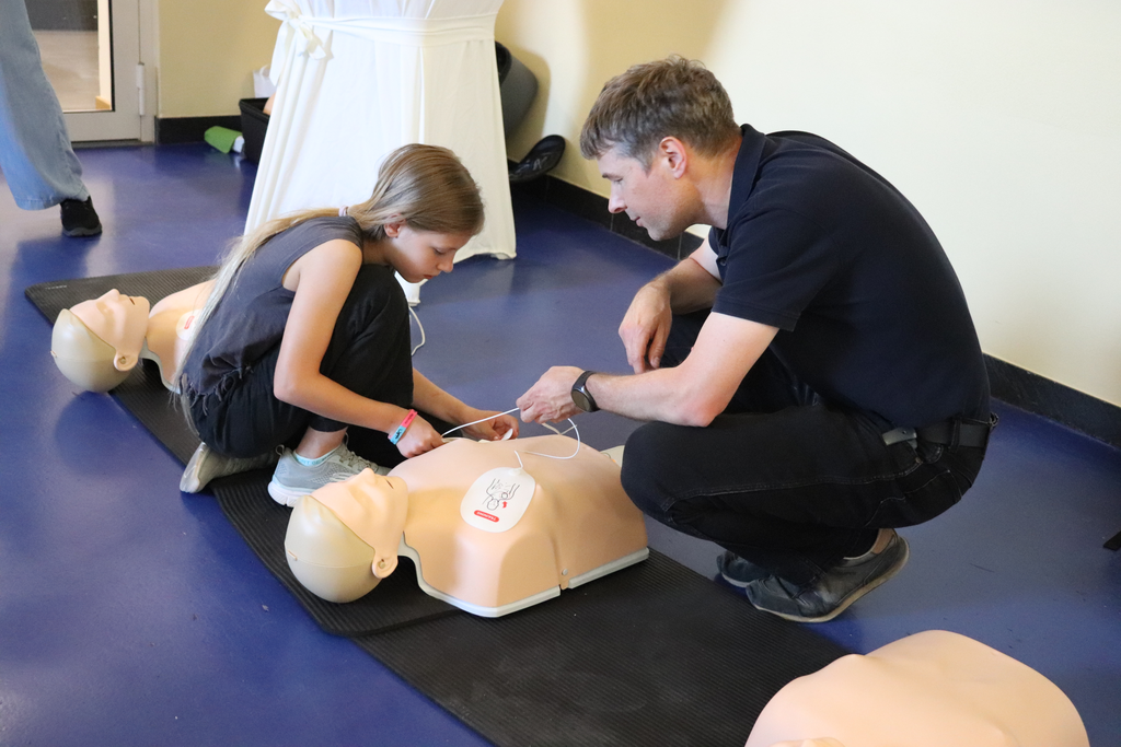 Ein Mädchen und ein Mann knien gemeinsam über einem Puppentorso und legen ihm einen Defibrillator an.